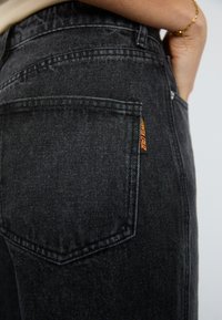 Jeans en denim noir avec une poche arrière portant une étiquette orange. Le matériau a une texture lisse et une finition légèrement délavée.