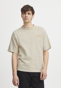Beige bomull t-skjorte med korte ermer, rund hals og en liten "CASUAL" logo i oransje-brun nyanse på brystet.