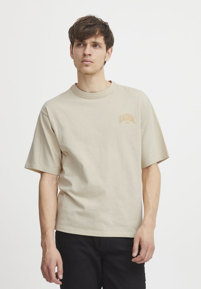 Beige bomull t-skjorte med korte ermer, rund hals og en liten "CASUAL" logo i oransje-brun nyanse på brystet.