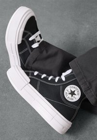 Sneaker in tela nera con suole in gomma bianca spessa, cuciture nere e lacci bianchi. Presenta una toppa con logo circolare sul lato.