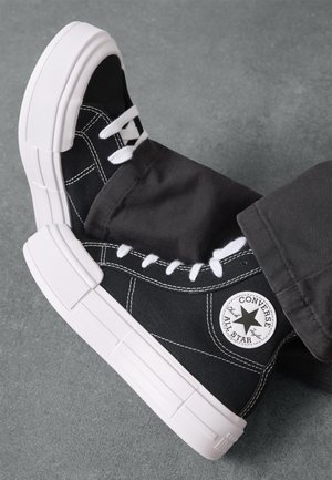 Baskets en toile noires avec semelles en caoutchouc blanc épaisses, coutures noires et lacets blancs. Présente un patch logo circulaire sur le côté.