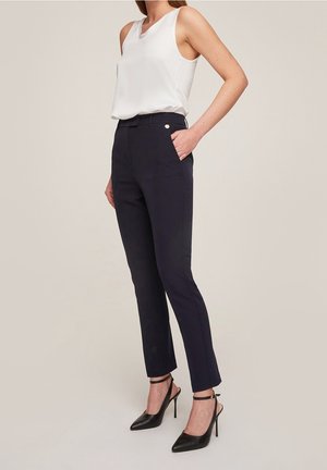 Femme portant un chemisier blanc sans manches, un pantalon ajusté bleu marine et des escarpins noirs à bout pointu avec brides à la cheville sur fond beige.