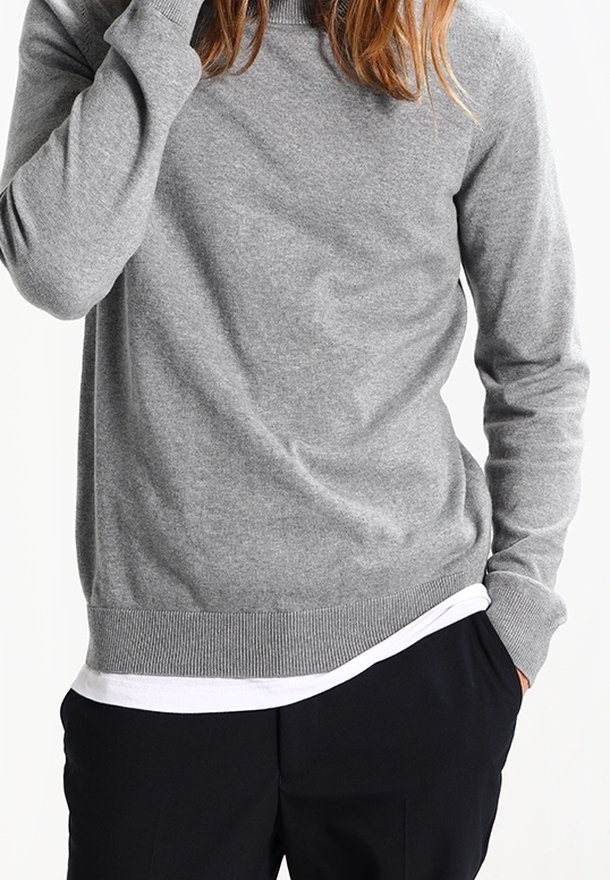 Personne portant un pull gris à manches longues sur une chemise blanche, une main dans la poche du pantalon noir et l'autre près du visage.