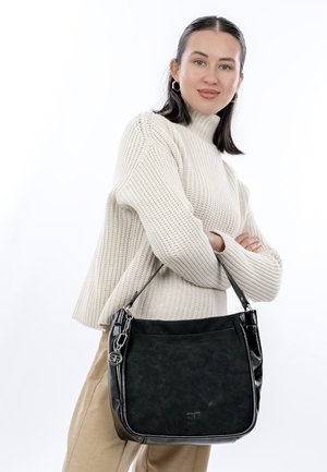 Moteris vilkinti smėlio spalvos megztinį ir kelnes, laiko juodą peties rankinę su sidabriniu pakabuku, pozuoja prieš baltą foną.