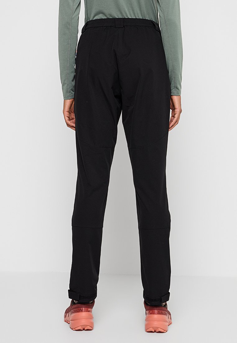 Czarne spodnie zewnętrzne o luźnym kroju, z elastycznym pasem i widocznymi detalami szwów. W połączeniu z czerwonawymi butami turystycznymi.