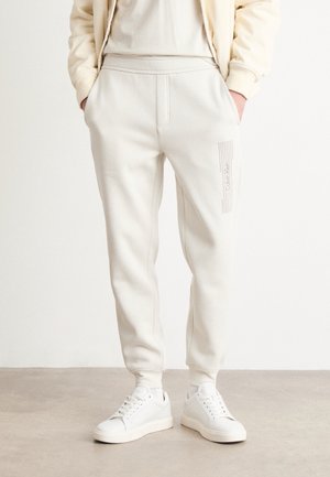 Person trägt off-white Jogginghosen mit seitlichem Logo und passende weiße Sneakers, steht mit den Händen in den Taschen vor einem einfarbigen Hintergrund.