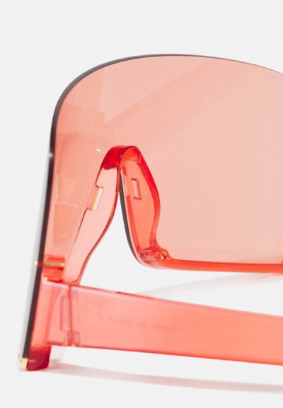 Gafas de sol tintadas en rojo con un marco transparente grueso, que presenta formas geométricas audaces y detalles en dorado en las bisagras.