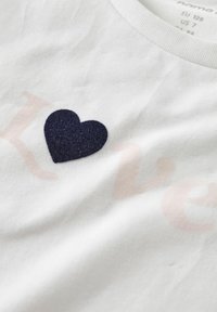 Valge T-särk, millel on tumesinise villase südame aplikaator ja osaliselt nähtav helesinine kiri. Pehme kangas sileda tekstuuriga.