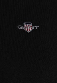 Schwarzer Stoff mit gerippter Textur und einem kleinen gestickten Logo in Grau und Rot, das ein Schilddesign und den Text "GANT" zeigt.