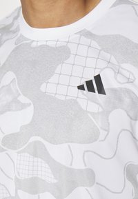 Zbliżenie na osobę w białej i szarej koszuli w wzory z czarnym logo Adidasa na piersi.