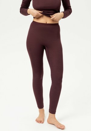 Persoon draagt aansluitende donkerpaarse legging en een bijpassende top met lange mouwen, tilt de top op om de blote buik te laten zien, staat blootsvoets op een witte vloer.