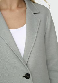 Lichtgrijs blazer van een gestructureerde stof, met een notch-lapel en een enkele zwarte knopen sluiting. Draagt over een wit top.
