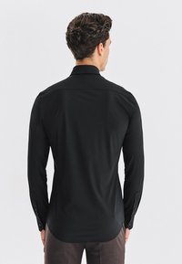 Czarna koszula z długim rękawem z gładkiego materiału, z zaokrąglonym dołem, kołnierzykiem i mankietami na guziki. Widok od tyłu ukazuje dopasowany krój.