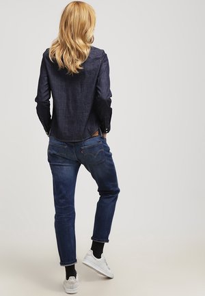 Femme aux cheveux blonds portant une chemise en jean foncé, un jean bleu, des chaussettes noires et des baskets blanches, debout de dos face au spectateur.