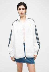 Νεαρός/ή φοράει μια άσπρη ζακέτα Adidas με μαύρες ρίγες και μια φούστα από τζιν, στέκεται μπροστά σε ένα απλό ανοιχτό φόντο.