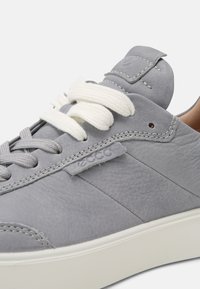 Sneaker grigio in pelle con lacci in cotone bianchi, dettagli cuciti e un discreto patch del logo. Presenta una superficie testurizzata e una suola leggera.