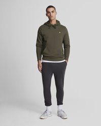 Sweat à capuche vert avec un cordon de serrage et une poche avant, associé à un pantalon de survêtement gris. La tenue présente un logo jaune et des baskets blanches.