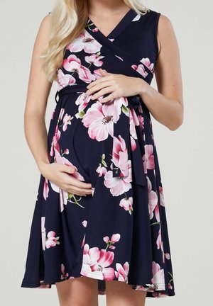 Schwangere Frau in einem marineblauen Blumenmuster-Kleid mit rosa und weißen Blumen, die sanft ihren Babybauch mit beiden Händen hält.