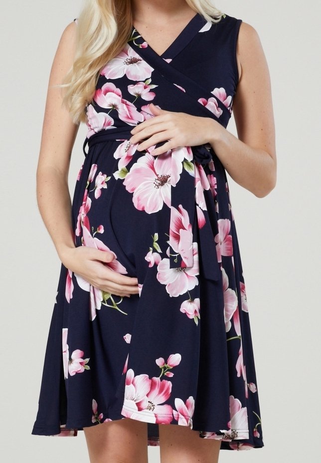 Gravid kvinna klädd i en marinblå blommig klänning med rosa och vita blommor, varsamt kramandes sin gravidmag med båda händerna.