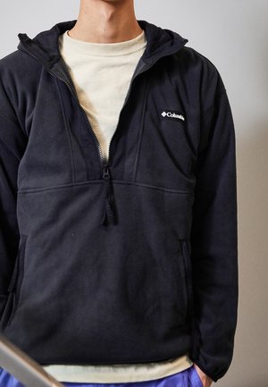 Mann trägt eine offene schwarze Columbia Fleecejacke über einem weißen T-Shirt, mit den Händen in den Taschen vor neutralem Hintergrund.