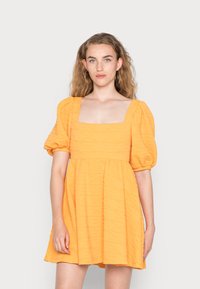 Vestido curto com mangas bufantes em tecido amarelo texturizado; decote quadrado e saia rodada. Apresenta um padrão subtil em todo o material.