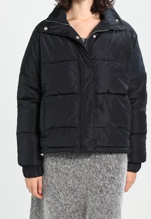 Veste matelassée noire à col montant et fermeture éclair à l'avant portée sur une jupe en maille grise, vue des épaules jusqu'à mi-cuisse.