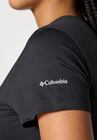 Nærbilde av en person som har på seg en svart kortermet skjorte med den hvite Columbia-logoen på ermene.