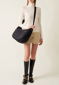 Sac à épaule noir texturé avec une fermeture éclair dorée et une sangle contrastante. Modèle portant un col roulé crème, un short beige et des bottes noires hautes.