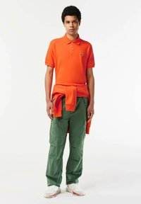 Camisa polo naranja brillante con cuello, combinada con pantalones cargo verdes con bolsillos laterales. Zapatillas blancas con acentos rojos completan el look.