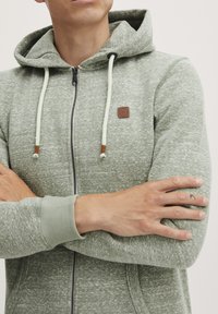 Haut à capuche zippé vert clair en tissu texturé. Il comporte un cordon gris, un écusson en cuir et des poignets côtelés.