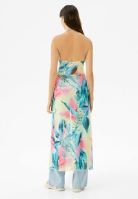 Vrouw met lang bruin haar, gekleed in een rugloze bloemen halterjurk over lichtblauwe jeans en crèmekleurige schoenen, staande met de rug naar de camera op een witte achtergrond.