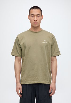 Un homme portant un T-shirt Snow Peak vert olive et un pantalon foncé se tient devant un fond blanc uni.