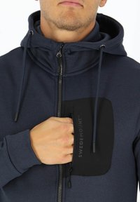 Felpa con zip di colore navy, dotata di una tasca sul petto nera testurizzata. Presenta un cappuccio regolabile con cordino e chiusura frontale con zip. Tessuto morbido al tatto.