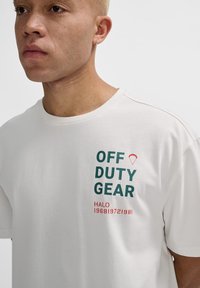 Vit bomull t-shirt med texten "OFF DUTY GEAR" i grön och röd färg, korta ärmar och en klassisk rund halsringning.