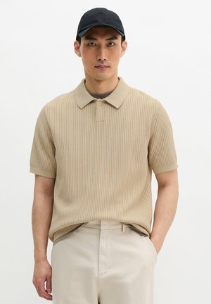 Mann trägt beiges, strukturiertes kurzärmliges Polohemd, hellbeige Hose und schwarze Kappe, steht mit einer Hand in der Tasche vor weißem Hintergrund.