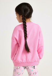 Vaaleanpunainen huppari, jossa on roikkuvat olkapäät ja ribbireunat; yhdistetty kevyisiin vaaleanpunaisiin leggingseihin, joissa on sarjakuvahahmoja ja tähtiä.