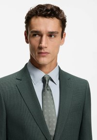 Grüne, strukturierte Anzugjacke mit einem Revers, kombiniert mit einem hellblauen Hemd mit Kragen und einer grünen gemusterten Krawatte mit kleinen Quadraten.