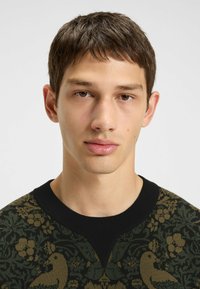 Un giovane con i capelli corti e castani indossa un maglione verde scuro e nero con un motivo floreale e di uccelli.