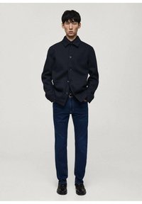 Mornarska jakna na duge rukave od teksturirane tkanine, u kombinaciji s tamnoplavim trapericama i crnim cipelama, prikazana na neutralnoj pozadini.