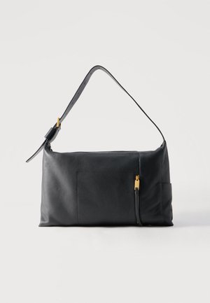 Mustast nahast õlakott reguleeritava rihma ja kuldse luku detailiga esitaskul, lihtsa hele tausta ees.