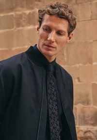 Giacca bomber nera con chiusura a zip, abbinata a una cravatta a fantasia paisley in toni scuri, indossata su una camicia con colletto. Tessuto texturizzato.