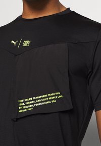 Svart sporttröja med en texturerad panel. Har en bröstficka och ljusgröna texter som säger "FÖRSTA MILEN" samt ett återvinningsbudskap.