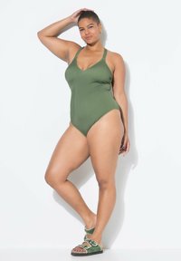 Olivově zelené jednodílné plavky s hlubokým V-výstřihem, texturovanými ramínky a přiléhavým střihem, v kombinaci se zelenými plochými sandály s dekorativními prvky.