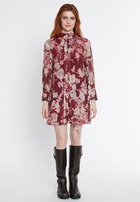 Langärmliges Kleid mit tiefrotem Blumenmuster, aus transparentem Stoff mit hohem Kragen und plissierten Details. Kombiniert mit knielangen schwarzen Stiefeln.