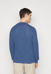 Ronde hals, lange mouwen trui van gestructureerde marineblauwe stof, met ribbels aan de mouwen en onderkant. Van achteren weergegeven, gecombineerd met lichte broeken.