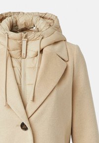 Cappotto trapuntato beige, staccabile, con una texture liscia, caratterizzato da una chiusura con zip, cappuccio con cordino e un singolo bottone al colletto.