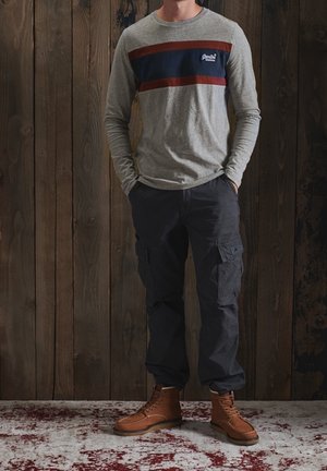 Grijze lange mouwen shirt met kastanjebruine en marineblauwe strepen, gecombineerd met donkere cargobroeken en bruine veterschoenen. Houten achtergrond.