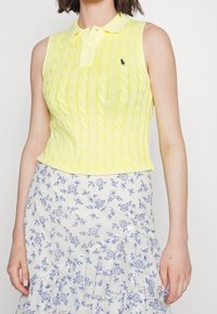 Žlutý pletený bezrukávový polo s kabelovým vzorem a límečkem. V kombinaci s bílou sukní s modrými květinovými vzory a volánkovým lemem.