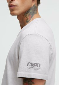 Helles graues Baumwoll-T-Shirt, kurze Ärmel, Rundhalsausschnitt, mit einem Logo an linken Ärmel und einer glatten, strukturierten Oberfläche.