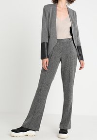 Kvinna som bär en grå kavaj med svarta lädermanchetter, matchande högmidjade byxor, beige ribbad linne och kraftiga svartvita sneakers.